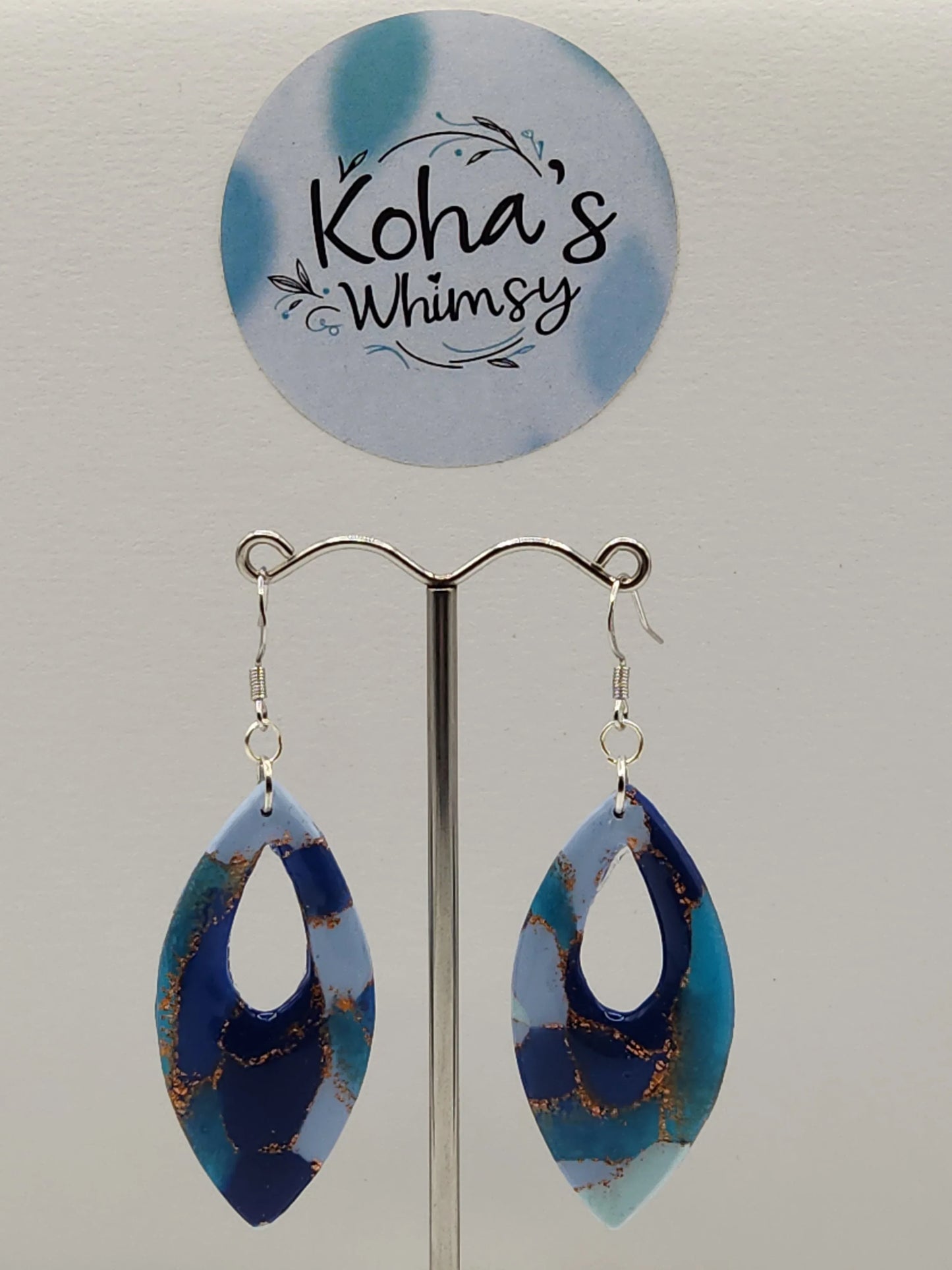 Blue and teal dangle earrings by Koha's Whimsy with gold-colored inclusions, suspended from a silver-colored metal display.