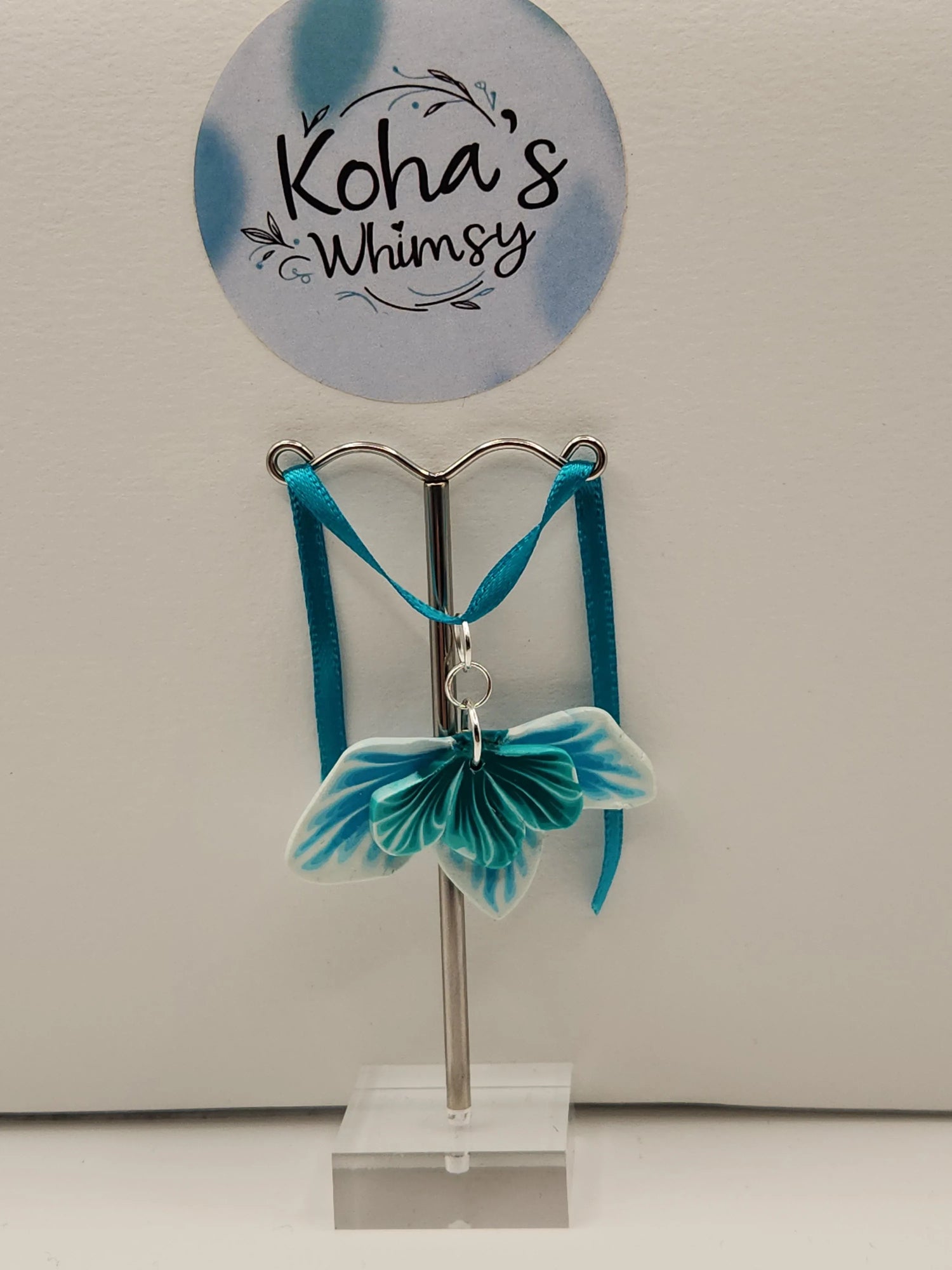 Teal leaf pendant by Koha's Whimsy with a white and blue swirl design, displayed on a small silver stand.
