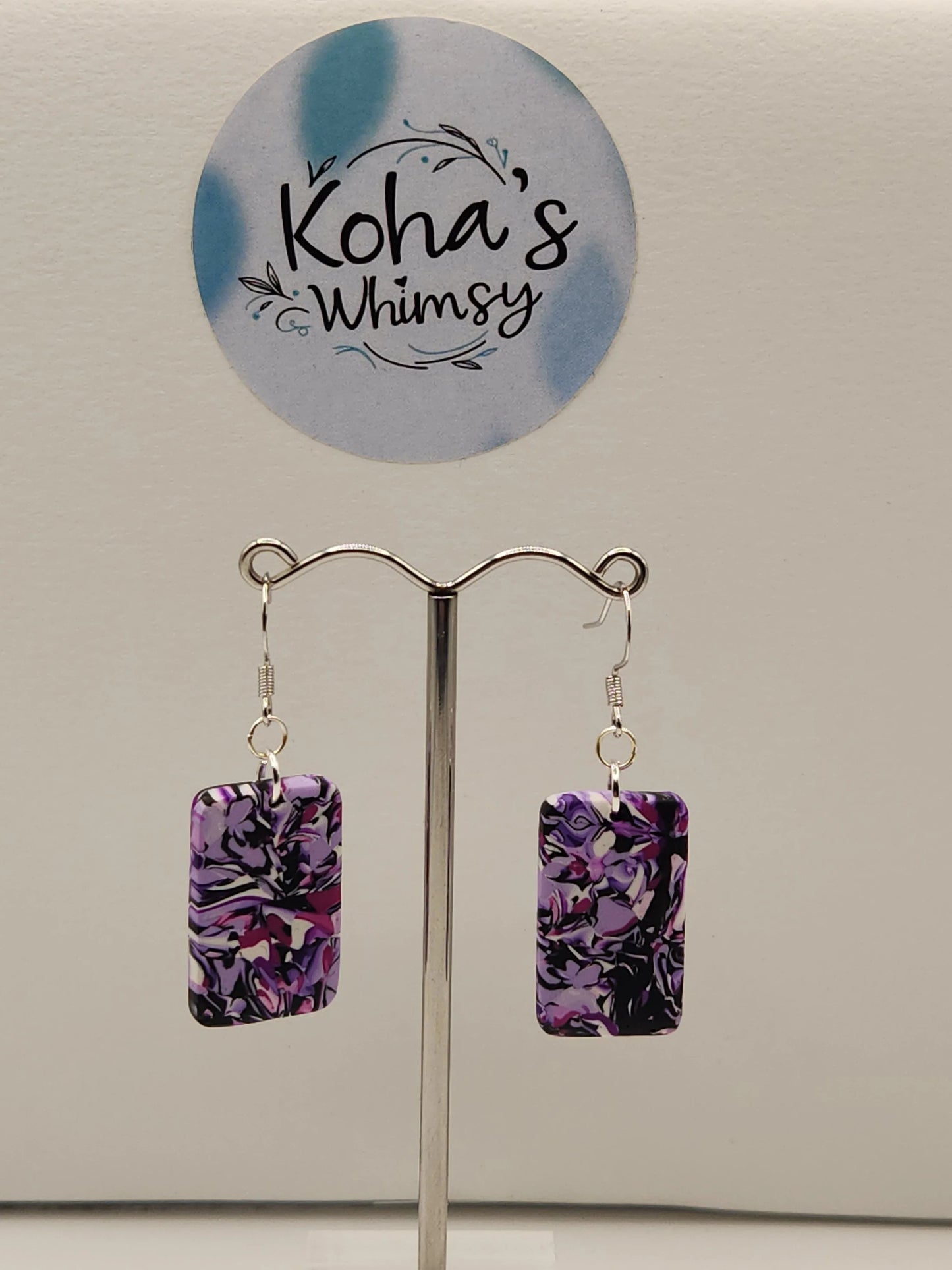 Rectangular dangle earrings in purple chaos print by Koha's Whimsy on a silver display stand.