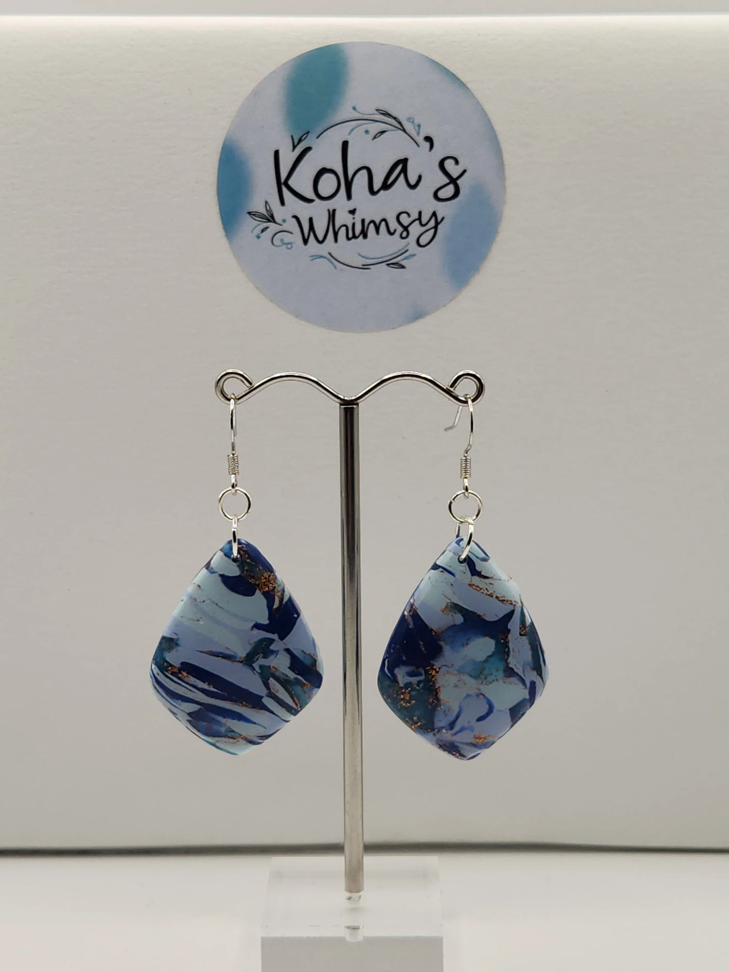 Blue diamond dangle earrings by Koha's Whimsy with gold detailing, hanging from a silver earring stand.