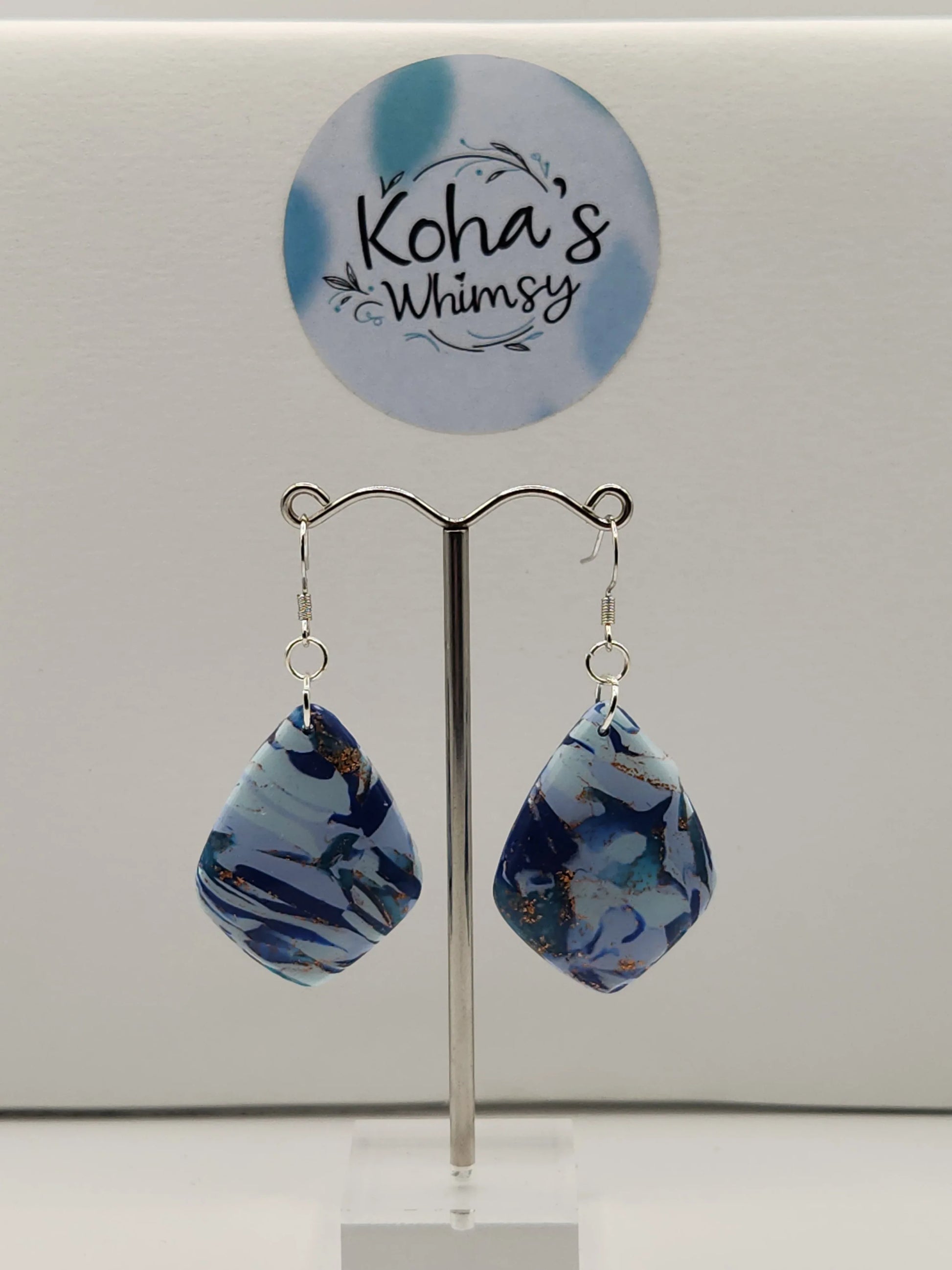 Blue diamond dangle earrings by Koha's Whimsy with gold detailing, hanging from a silver earring stand.